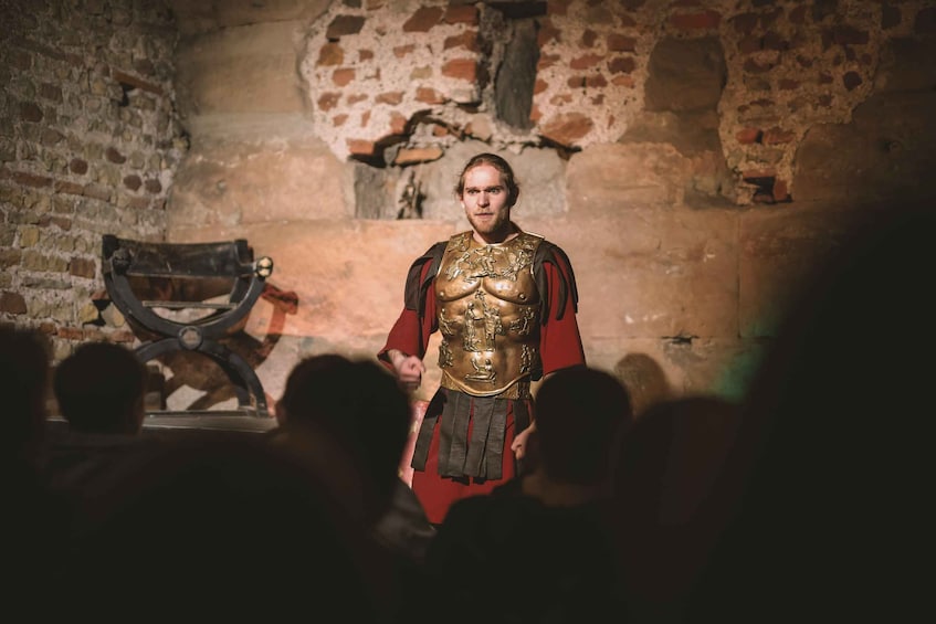 A person in historical armor standing in a stone-walled room with a wooden chair and a statue in the background. 5