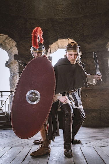 Two individuals in historical costumes, one holding a shield and the other a sword, standing in a stone structure. 3