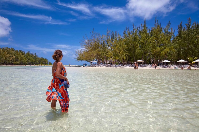 A woman in a colorful dress walking in shallow water, a beach with umbrellas, and a line of trees in the background. 4