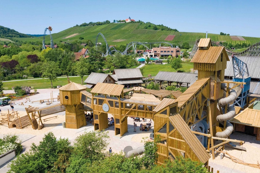 A wooden play structure with slides and climbing areas, set against a backdrop of green hills and a clear sky. 5