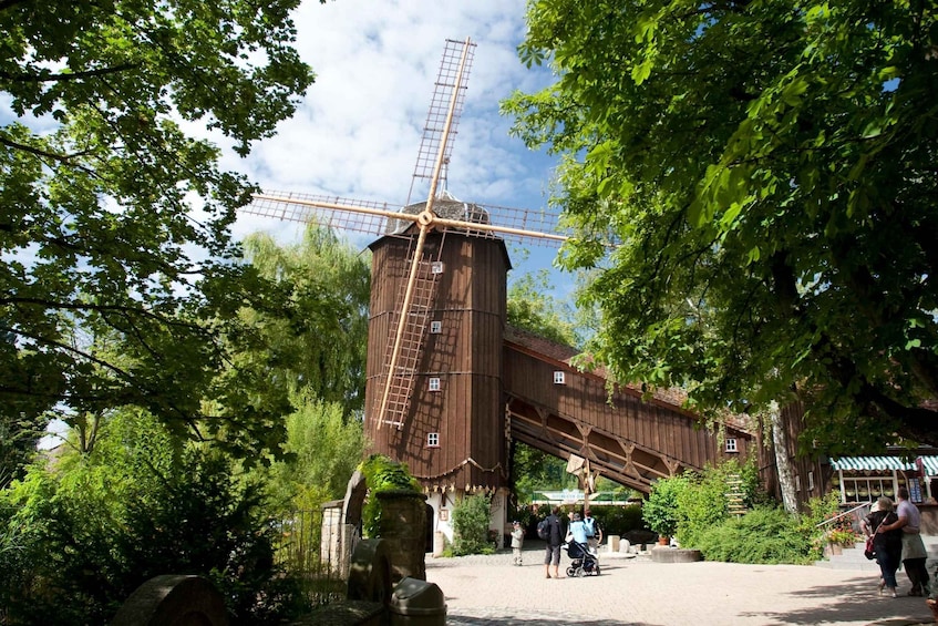 A wooden windmill with a ladder, surrounded by lush green trees and a paved area with people. 2