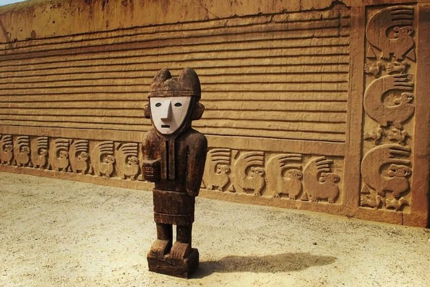 Una figura de piedra tallada con rostro similar a una máscara, ubicada frente a un muro con grabados similares. 2