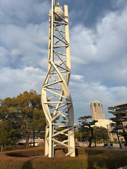 A tall, spiraling metal sculpture in a park with trees and buildings in the background. 2