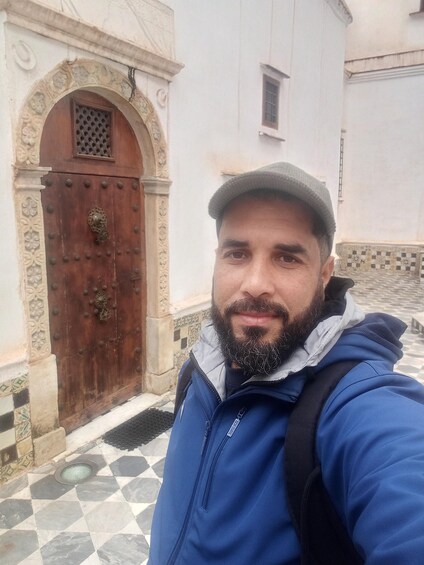 A man in a blue jacket and cap standing in front of a historic building with a decorative wooden door and a tiled entrance. 3