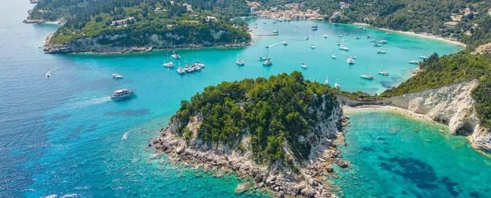 A coastal landscape with clear turquoise waters, a small rocky island, and numerous boats anchored in the bay. 4