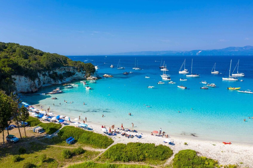A coastal beach with clear turquoise waters, numerous sailboats, and a sandy shore with beach umbrellas and people. 5