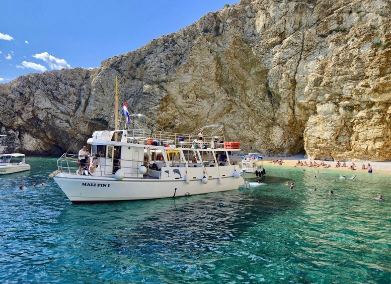 A boat named MALIPIN I on clear turquoise water with people swimming and sunbathing on the beach in the background. 1
