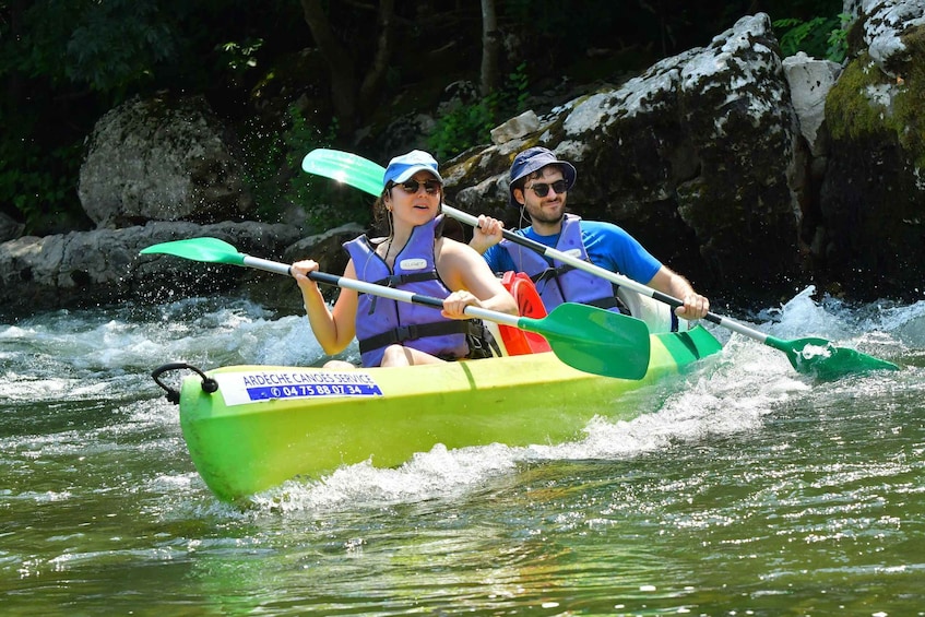 Two people kayaking on a river surrounded by rocks and greenery. 1
