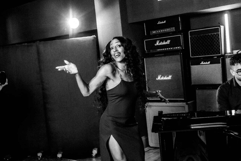 A person in a dark dress with a high slit, gesturing with their hand, standing in front of a Marshall amplifier. 4