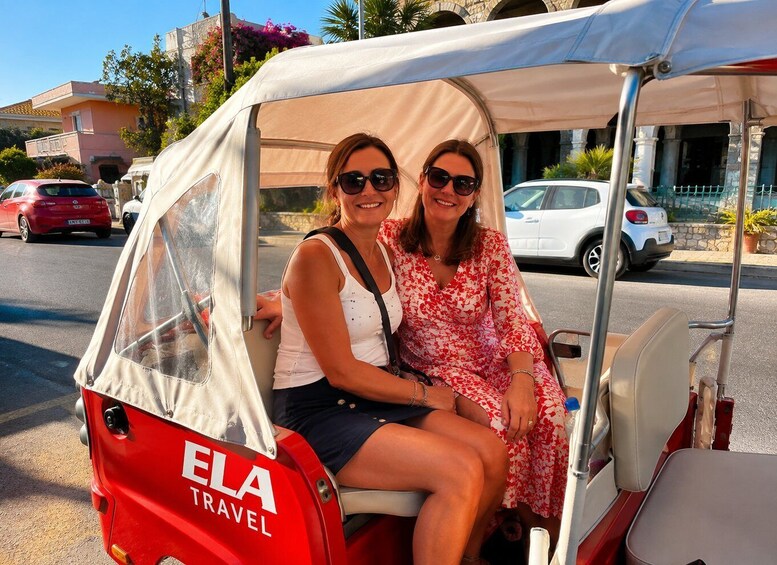 Two people in a red and white covered vehicle labeled 'ELA TRAVEL' on a street with parked cars and buildings. 1