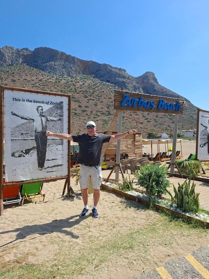 A man standing in front of a beach sign with a mountainous backdrop. 5
