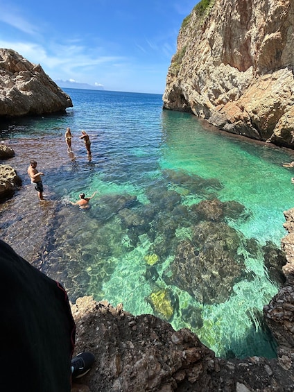 A rocky coastal area with clear turquoise water, people swimming and standing in the water, and a tent set up on the rocks. 3