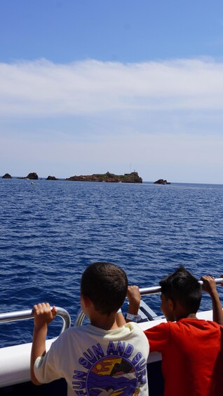 Two children on a boat, one wearing a white shirt with a graphic design, and the other in red, looking at a rocky island in the distance. 3
