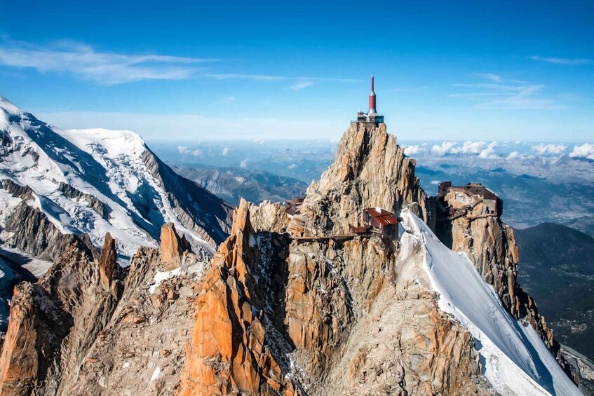 A rugged mountain peak with a red structure on top, snow-covered slopes, and a clear blue sky. 1
