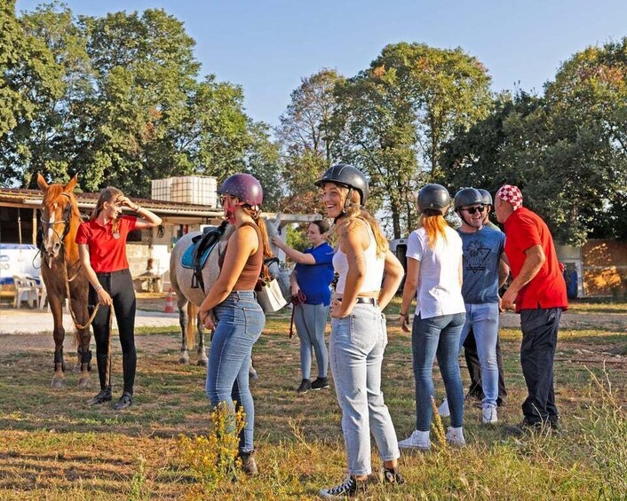 A group of people standing outdoors, some wearing helmets, with a horse and a building in the background. 5