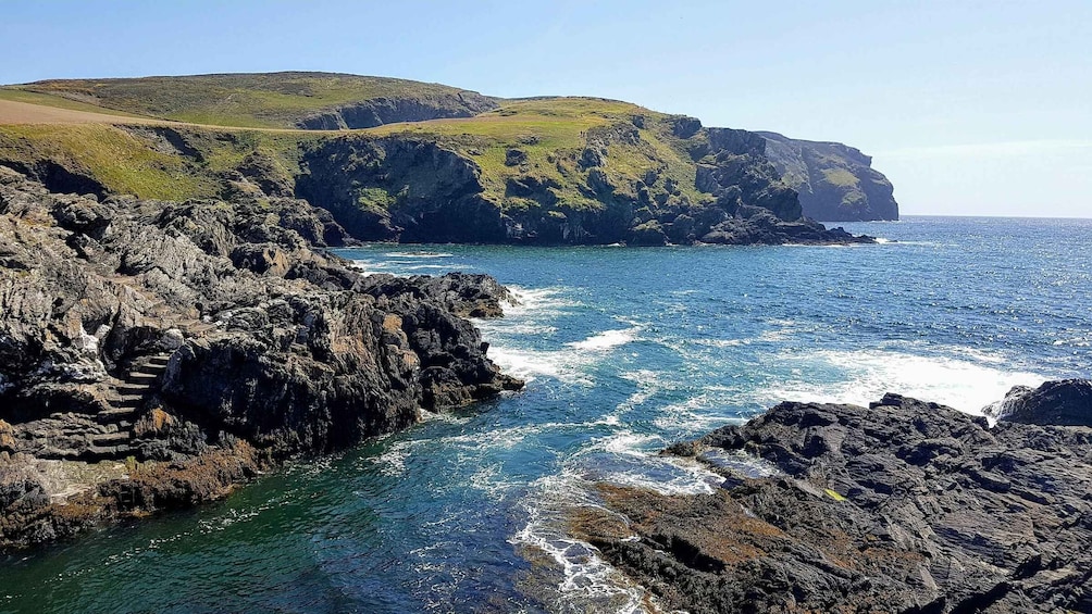 A coastal landscape with rocky cliffs, a clear blue sea, and a grassy hill. 5