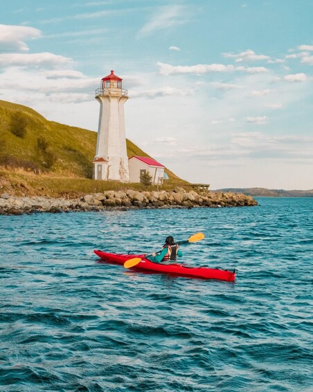 A lighthouse on a grassy hill by the sea, a red kayak with a person paddling, and calm blue waters. 4