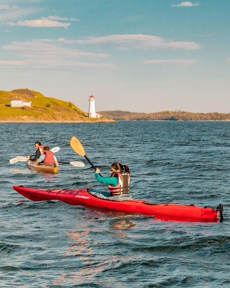 A red kayak with a person paddling, a lighthouse in the background, and a grassy hill with a white building. 2
