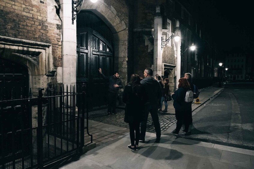 A group of people standing on a cobblestone street at night, with a brick building and a metal gate in the background. 2