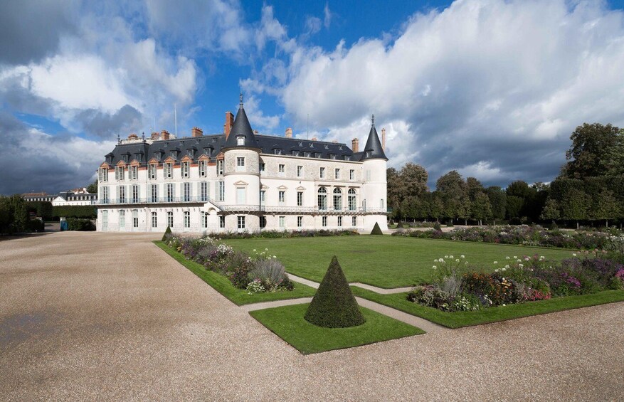 A large, historic building with a symmetrical facade, a central tower, and a well-manicured garden with various plants and a geometric lawn design. 3