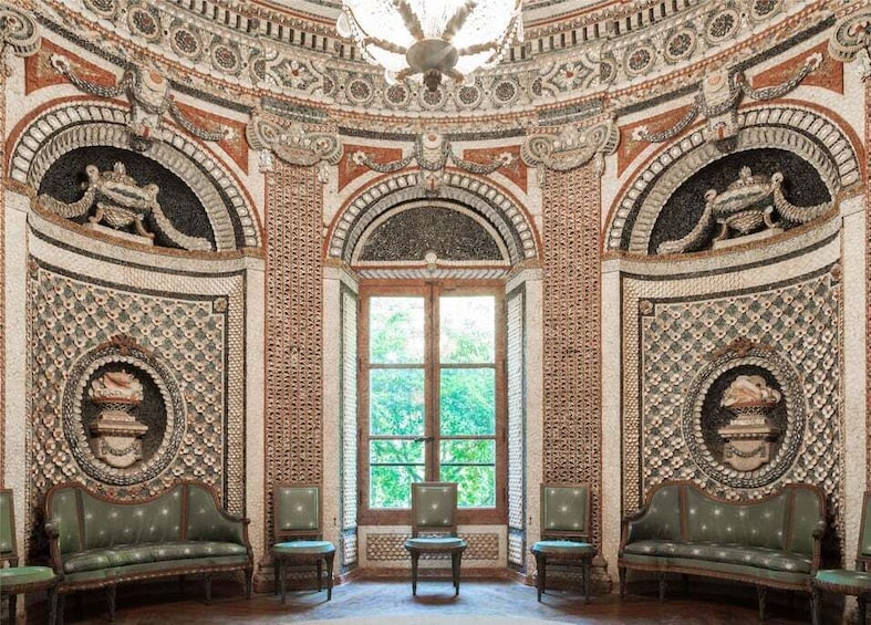 An ornate room with intricate mosaic walls, a large arched window, and green upholstered furniture. 1