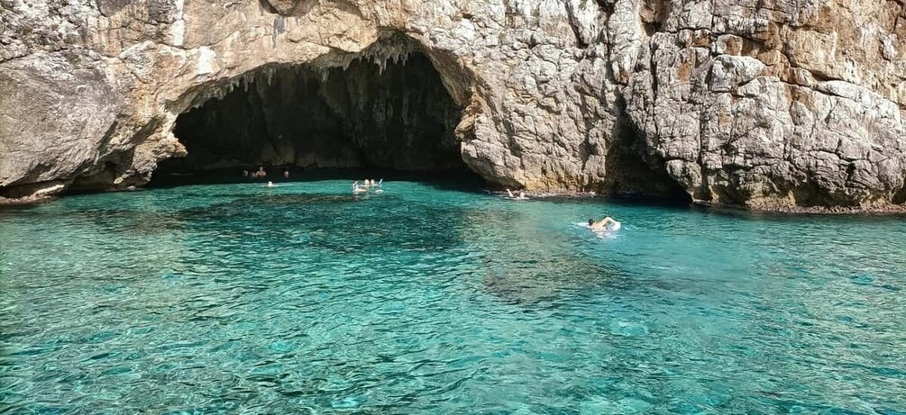 A clear turquoise sea with people swimming and a rocky cave entrance. 1