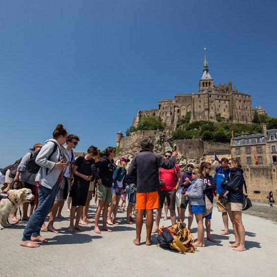 A group of people standing on a beach, facing a historic castle with a tower. 1