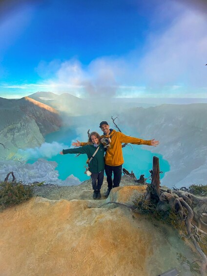 Two people standing on a mountain edge with arms outstretched, overlooking a valley with a turquoise lake and distant mountains. 1