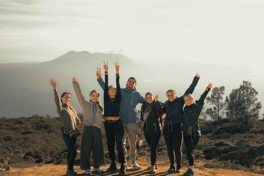 A group of people standing on a dirt path with their arms raised, facing a mountain range in the background. 2