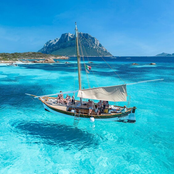 A sailboat with people on board in clear turquoise waters with a mountainous island in the background. 1
