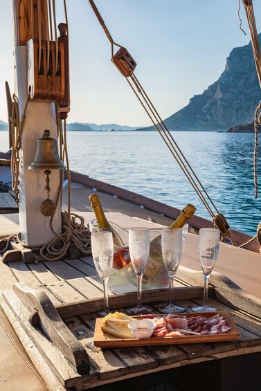 A boat deck with a table set for a meal, including a bottle of champagne, glasses, and a variety of meats. 3