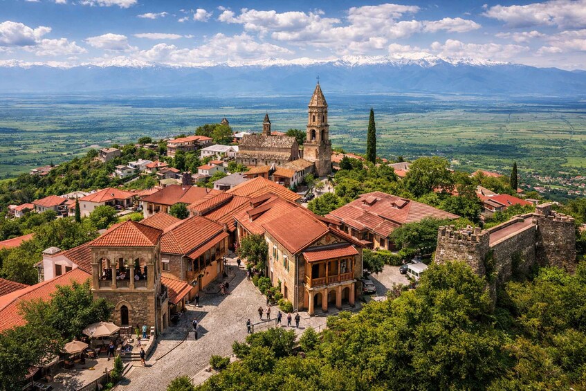 A historic town with red-roofed buildings, a prominent church with a bell tower, and a mountainous backdrop. 5