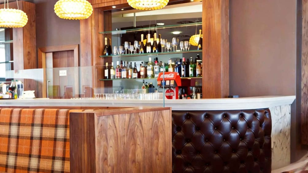 A bar with wooden shelves stocked with bottles and glasses, a red soda dispenser, and a tufted leather booth. 2