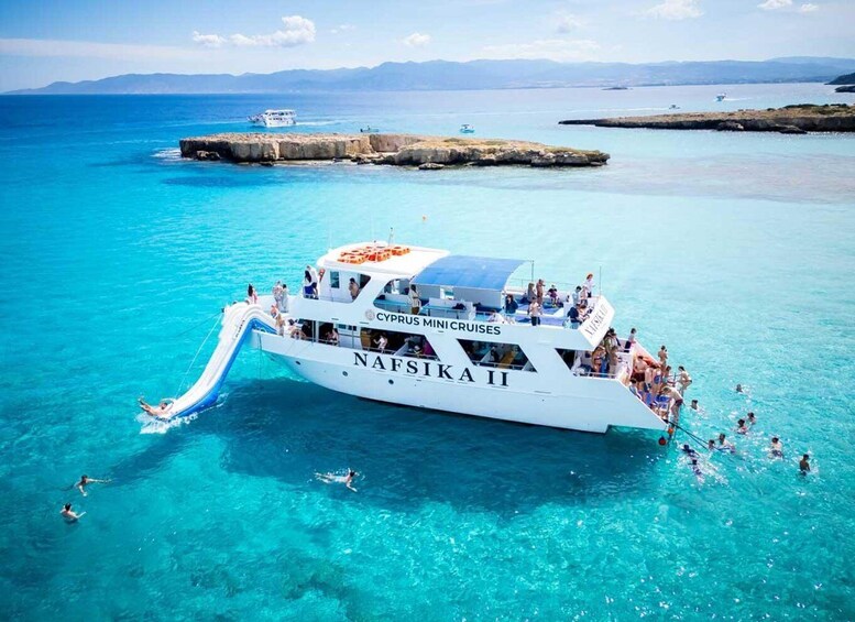 A white boat named 'NAFSIKA II' with 'CYPRUS MINI CRUISES' written on it, floating in clear turquoise waters with people on board and some swimming in the water. 4