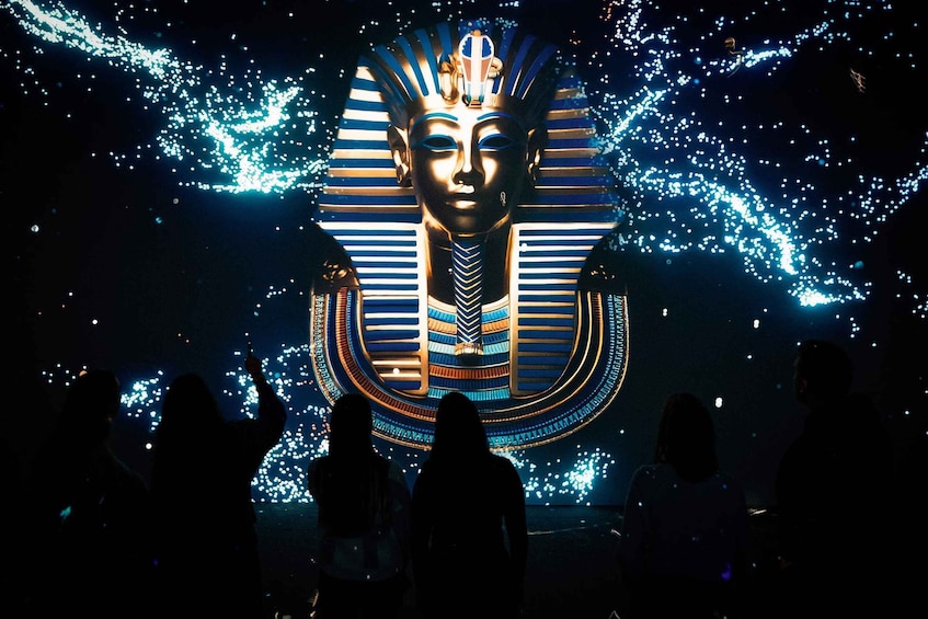A large illuminated pyramid structure with a face in the center, surrounded by people. 1