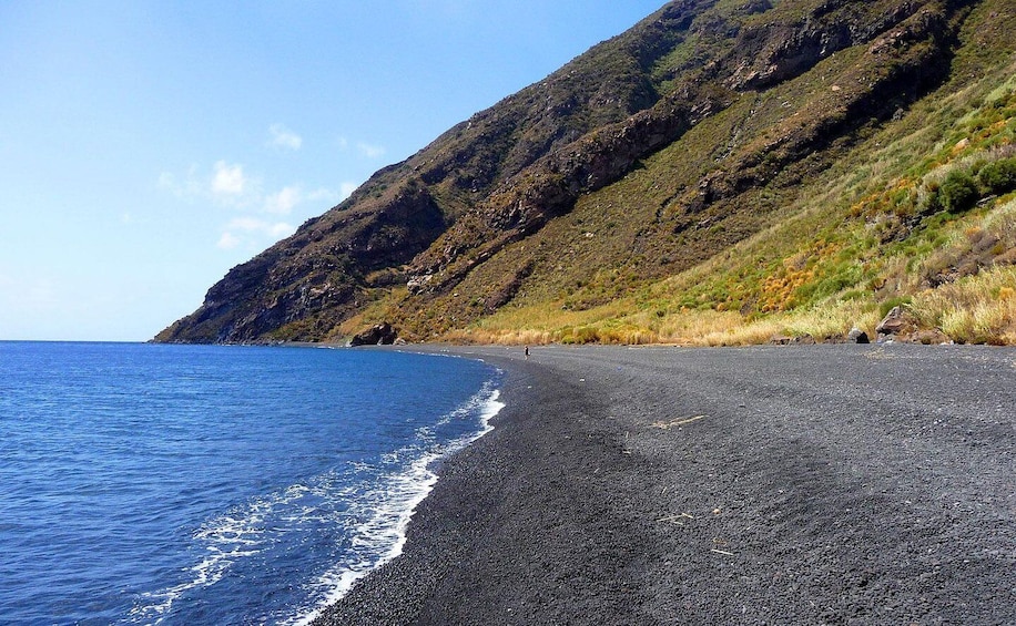 A black sand beach with a clear blue ocean and a steep, grassy hillside. 5