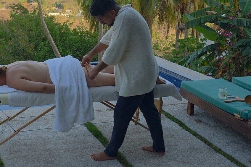 A person receiving a massage outdoors, with a pool and greenery in the background. 2
