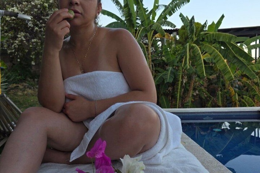 A person sitting by a pool with a white towel wrapped around them, surrounded by greenery and a clear blue sky. 5