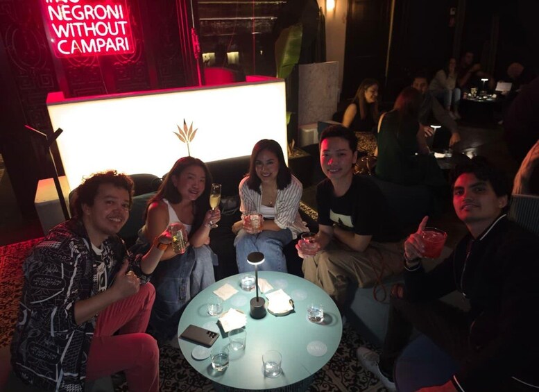 A group of people sitting around a table with drinks, in a dimly lit indoor setting with a neon sign in the background. 4