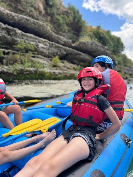 Two people in a blue raft wearing red life jackets and helmets, paddling on a river with rocky cliffs in the background. 4