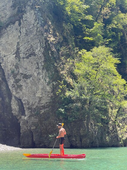 A person paddleboarding on a red board with a yellow paddle, surrounded by clear water and a rocky cliff with greenery. 4