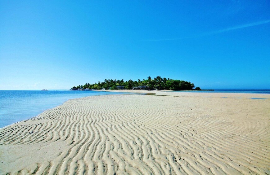 A sandy beach with gentle ripples, a clear blue sky, and a small island with trees in the distance. 2