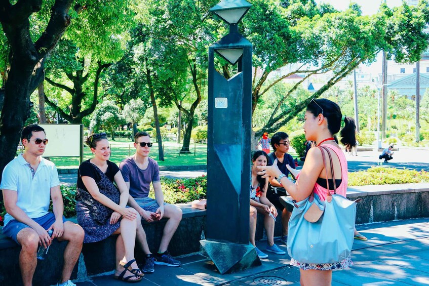 A group of people sitting on a bench in a park with trees and a tall, dark-colored lamp post. 2