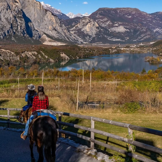 Two people riding horses on a road with a mountainous landscape and a lake in the background. 5