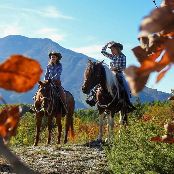 Two people riding horses on a trail with mountains in the background. 1