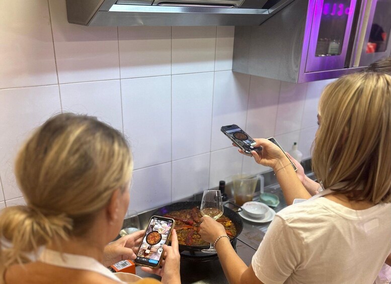 Two people using smartphones, one holding a glass of wine, in a kitchen with white tiles and a stainless steel range hood. 4