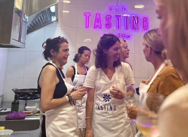 A group of people in a kitchen with a neon sign that reads 'ALC TASTING CLUB'. 5
