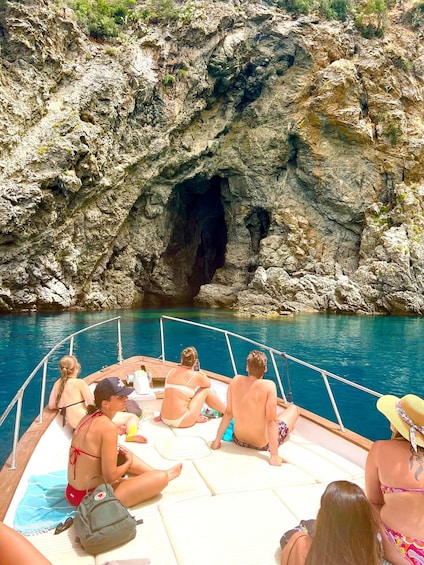 A boat with people sitting on the deck, a rocky cliff, and clear blue water. 2