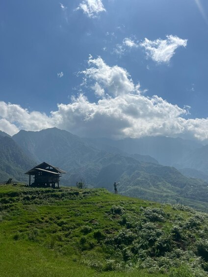 A mountainous landscape with a small wooden structure and a person standing on a grassy hill. 5