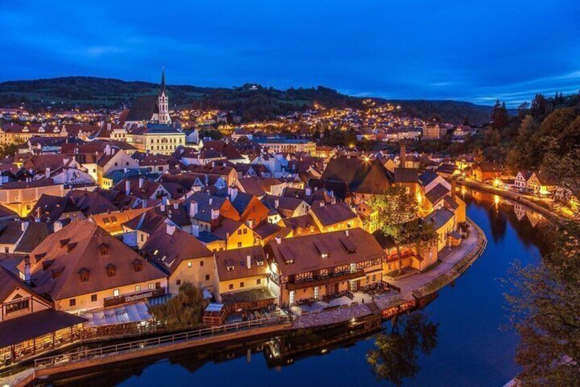 A town at dusk with illuminated buildings, a river, and a prominent church spire. 5